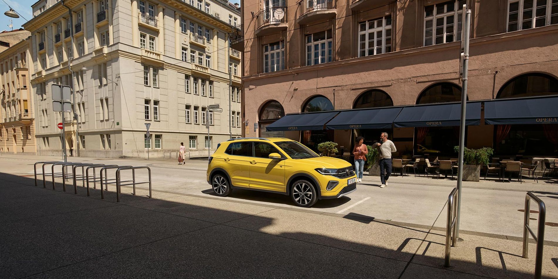 VW T-Cross in gelb parkt seitlich vor einem urbanen Straßencafé, ein Päarchen geht auf den Wagen zu.