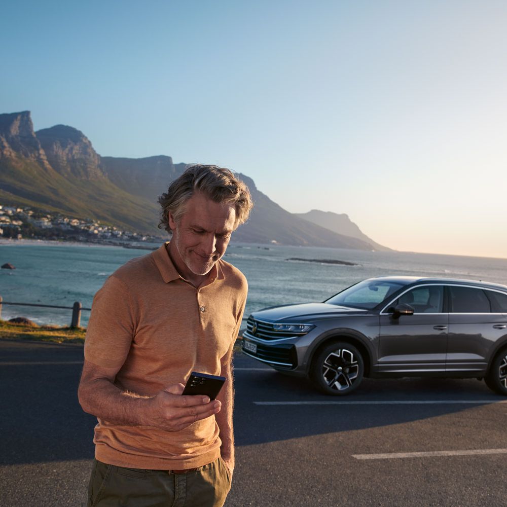 Ein Mann schaut auf sein Smartphone, im Hintergrund steht der Touareg vor einem See.