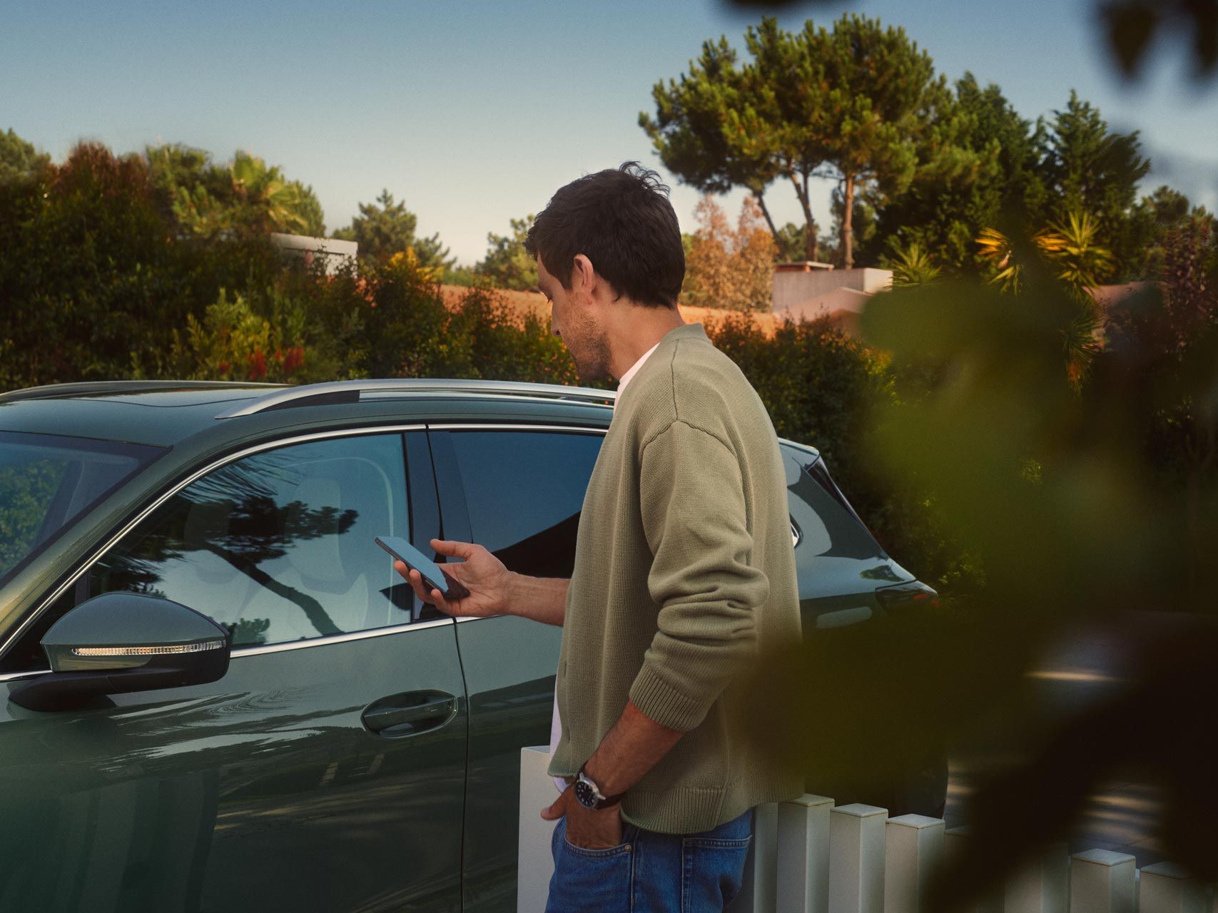 Ein junger Mann auf der Fahrerseite des dunkelgrünen VW Tiguan bedient den optionalen Park Assist Plus über sein Smartphone.
