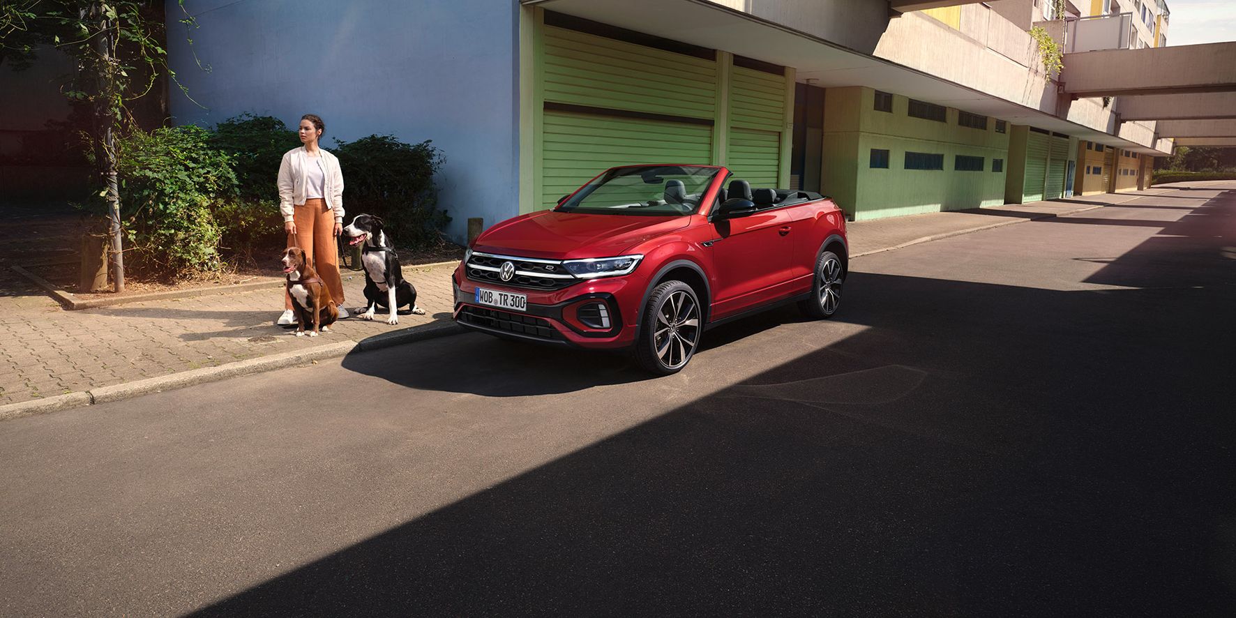 Rotes VW T-Roc Cabriolet mit geöffnetem Dach parkt am Straßenrand. Eine Frau mit zwei Hunden steht daneben. 