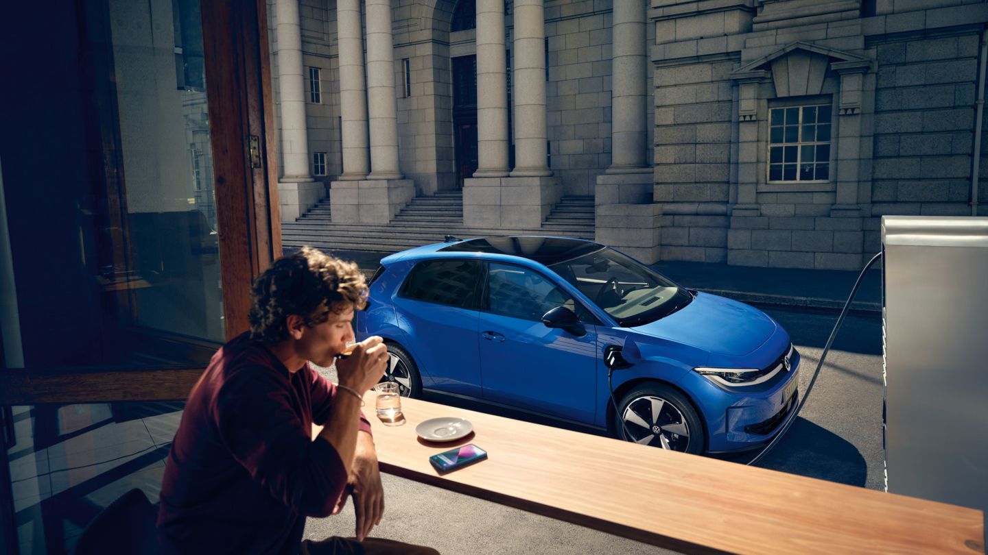 Egy férfi egy kávézóban ül, a nyitott ablakon át látható a kék VW ID. Polo, amint egy töltőállomásnál tölt.