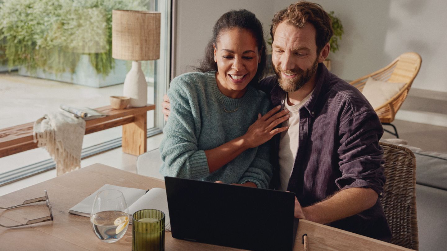 Egy pár mosolyogva ül egymás mellett egy faasztalnál, és együtt nézik a laptopot.