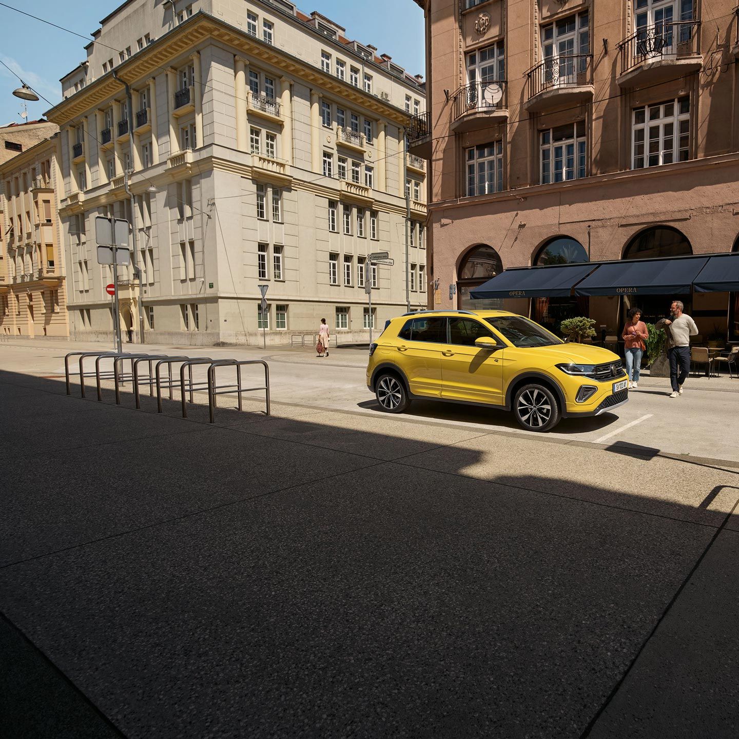VW T-Cross in gelb parkt seitlich vor einem urbanen Straßencafé, ein Päarchen geht auf den Wagen zu.