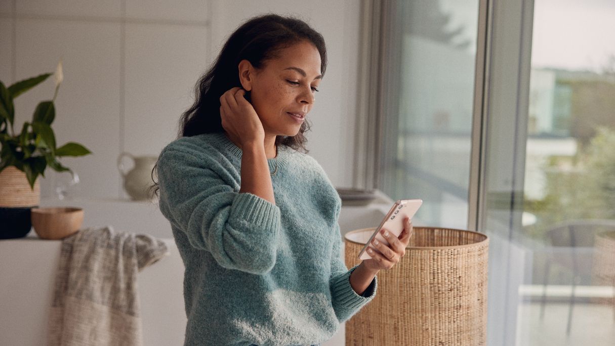 Egy nő otthon áll az ablaka előtt, és a mobiltelefonját nézi