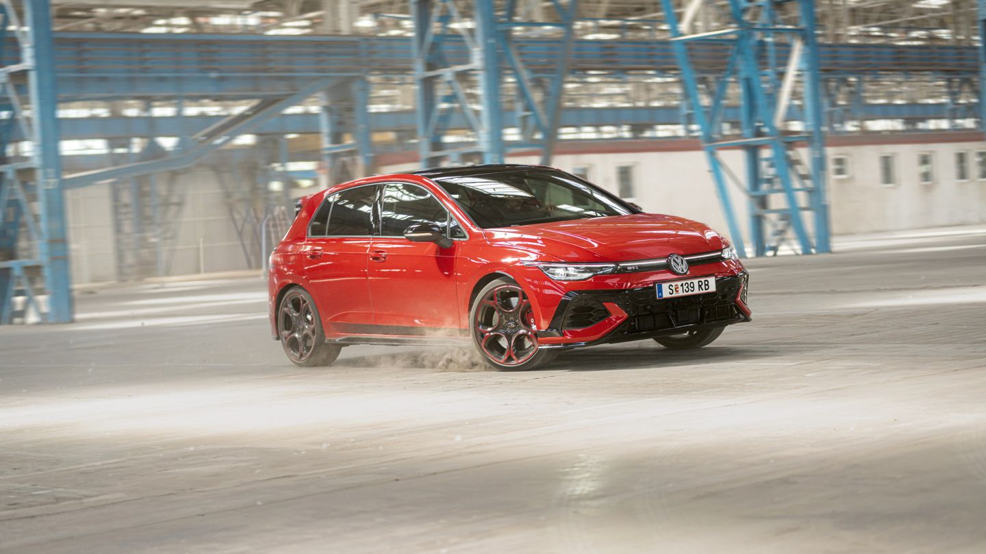 Piros Volkswagen Golf GTI halad gyorsan keresztül egy üres gyárépületben, miközben port kavar a kerekekkel.