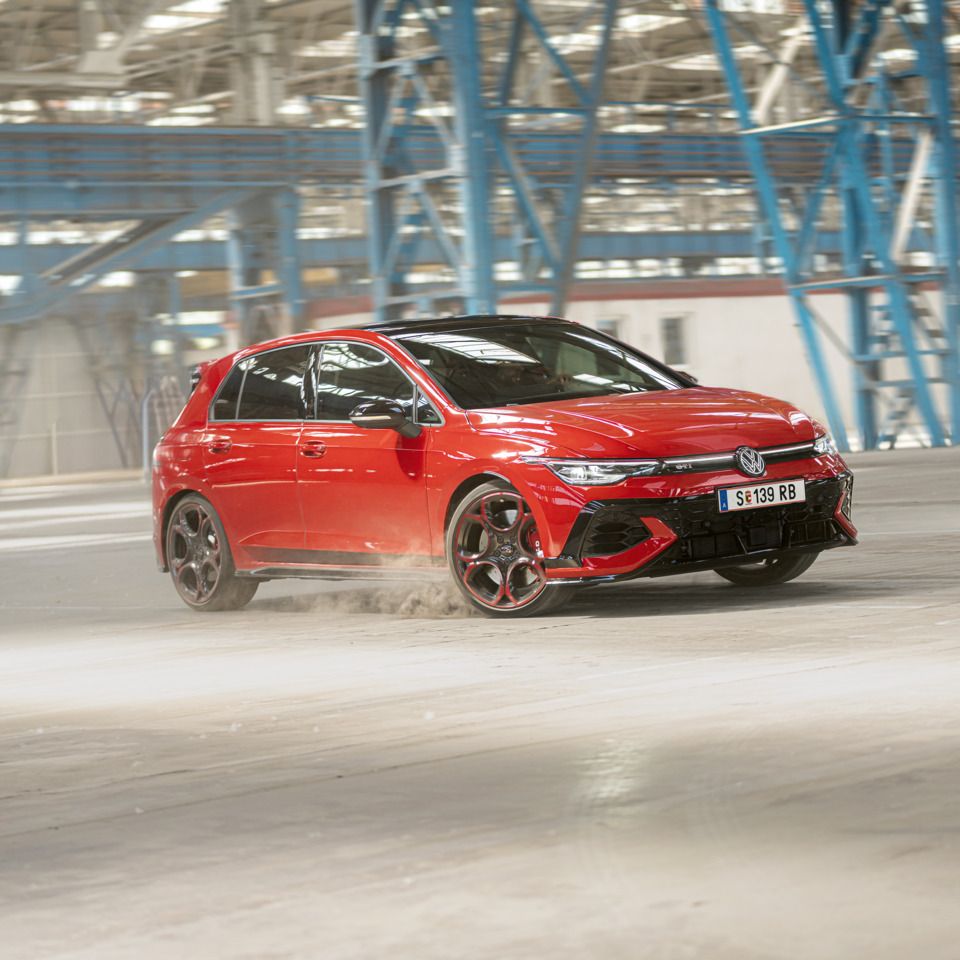 Piros Volkswagen Golf GTI halad gyorsan keresztül egy üres gyárépületben, miközben port kavar a kerekekkel.