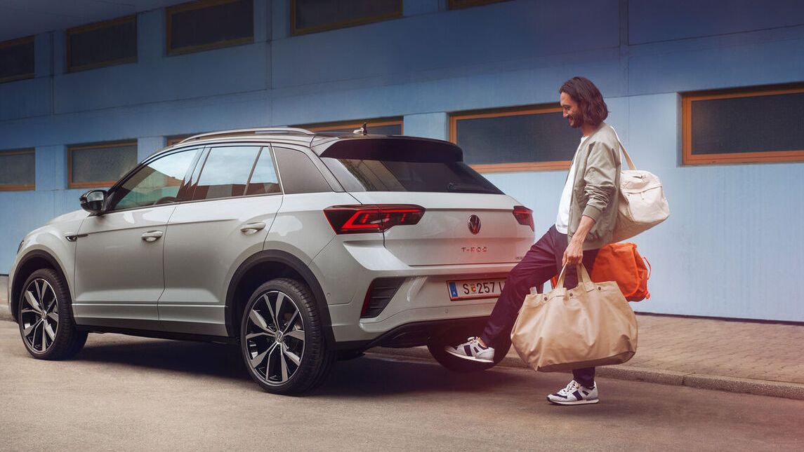 VW T-Roc R-Line in weiß vor einem Gebäude, Seite sichtbar, Mann steht hinter dem Auto und öffnet mit Fußschwenk die Kofferraumklappe