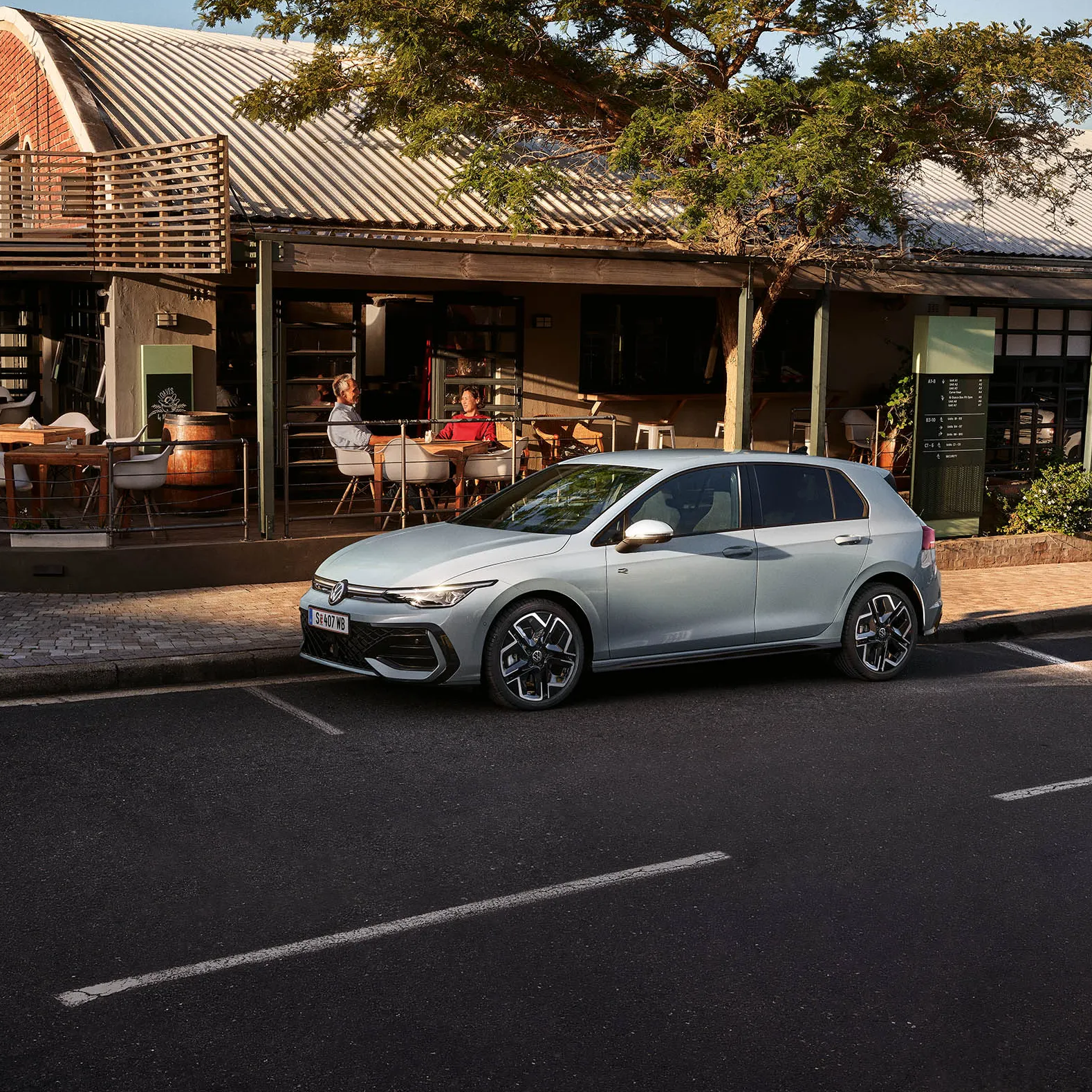 Bel Volkswagen Golf avtomobil je parkiran ob ulici pred restavracijo ali kavarno. Stavba ima leseno teraso za zunanjo postrežbo gostov. Na desni strani je parkiran še en bel avtomobil.
