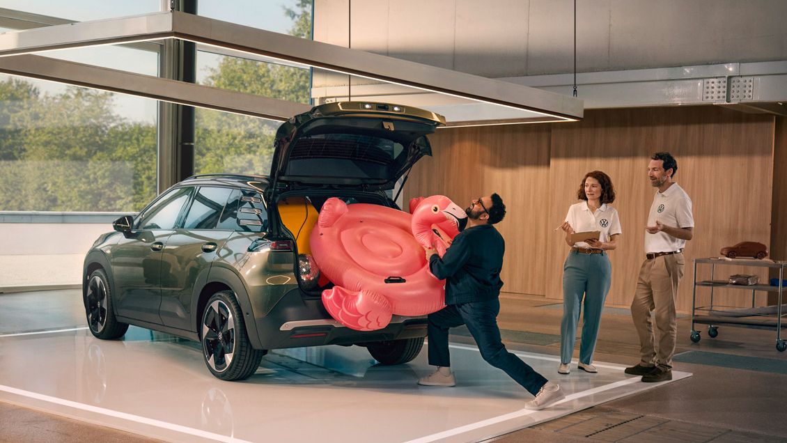 Egy férfi egy felfújható flamingót próbál betuszkolni a VW ID. Cross nyitott csomagterébe. Két személy áll mellette, és jót derülve figyeli.