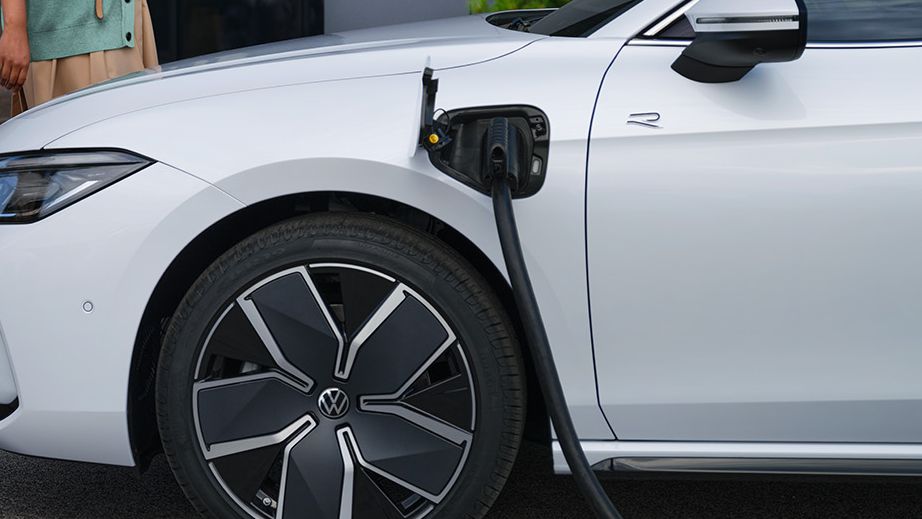 VW Passat fehér színben egy irodaház előtti oldalon, bedugva a töltőkábellel, két nő az autó felé sétál.