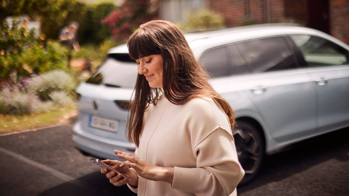  Egy nő nézi a telefonját, háttérben egy VW Golf Variant 