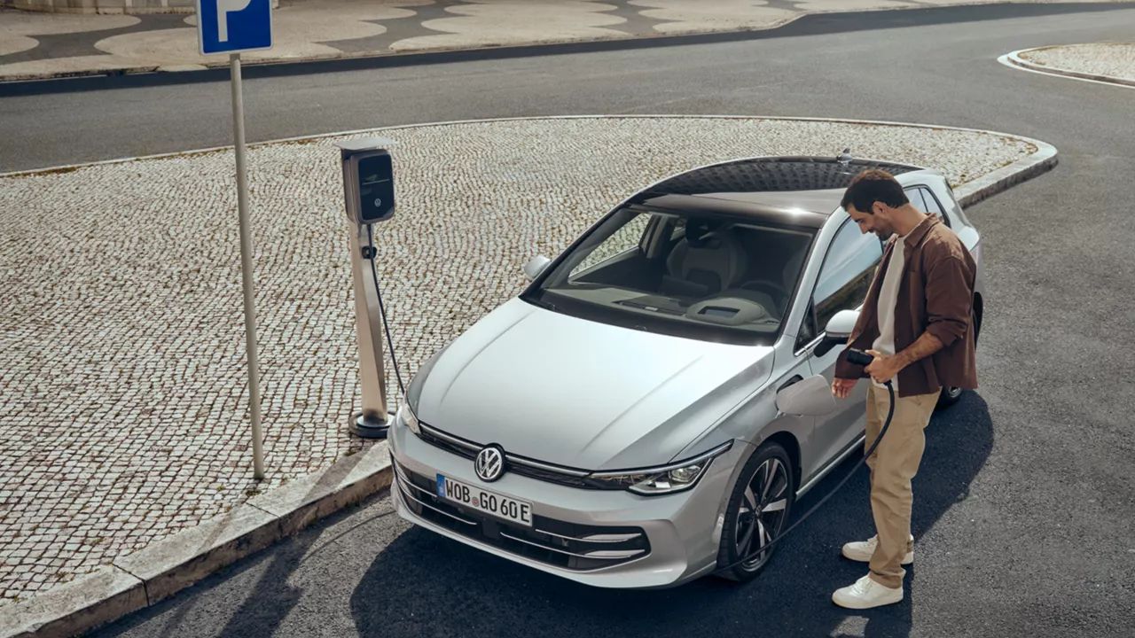 Egy ezüstszínű Volkswagen Golf Style plug-in hibrid áll egy városi parkoló töltőoszlopánál. Egy férfi áll az autó mellett, és kinyitja a töltőnyílás fedelét.