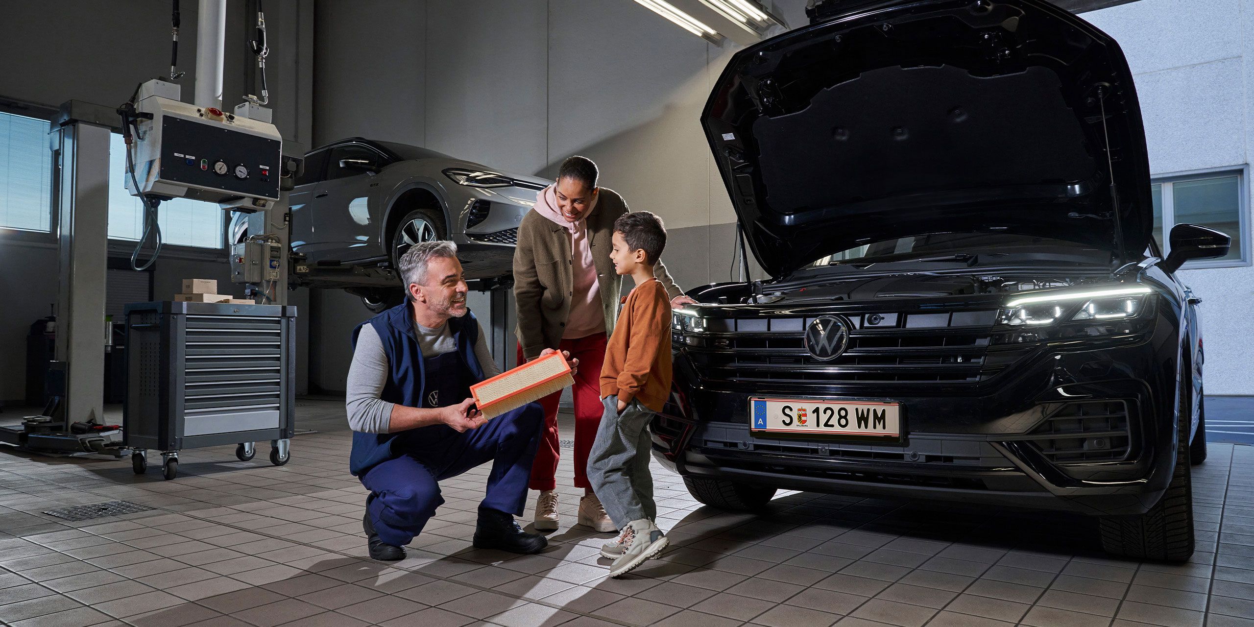 Ein Mechaniker erklärt den Aufbau eines Filters in einer Autowerkstatt. Im Hintergrund sind ein angehobenes Auto und ein geöffneter Motorraum eines schwarzen Touareg