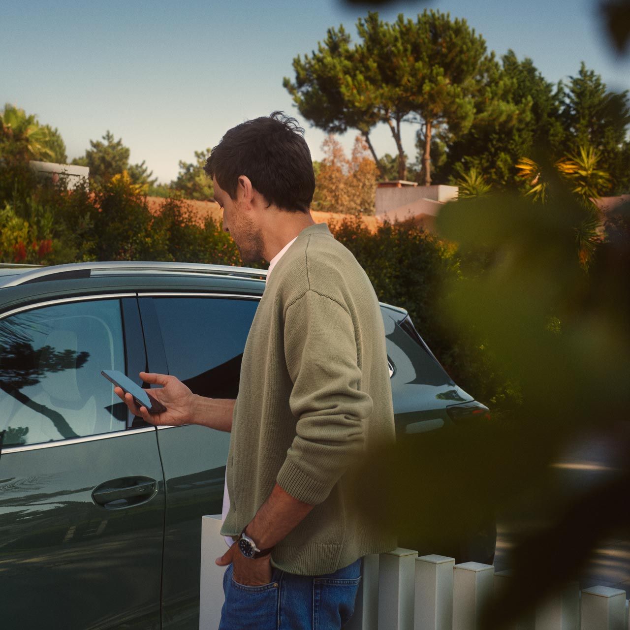 Ein junger Mann auf der Fahrerseite des dunkelgrünen VW Tiguan bedient den optionalen Park Assist Plus über sein Smartphone.