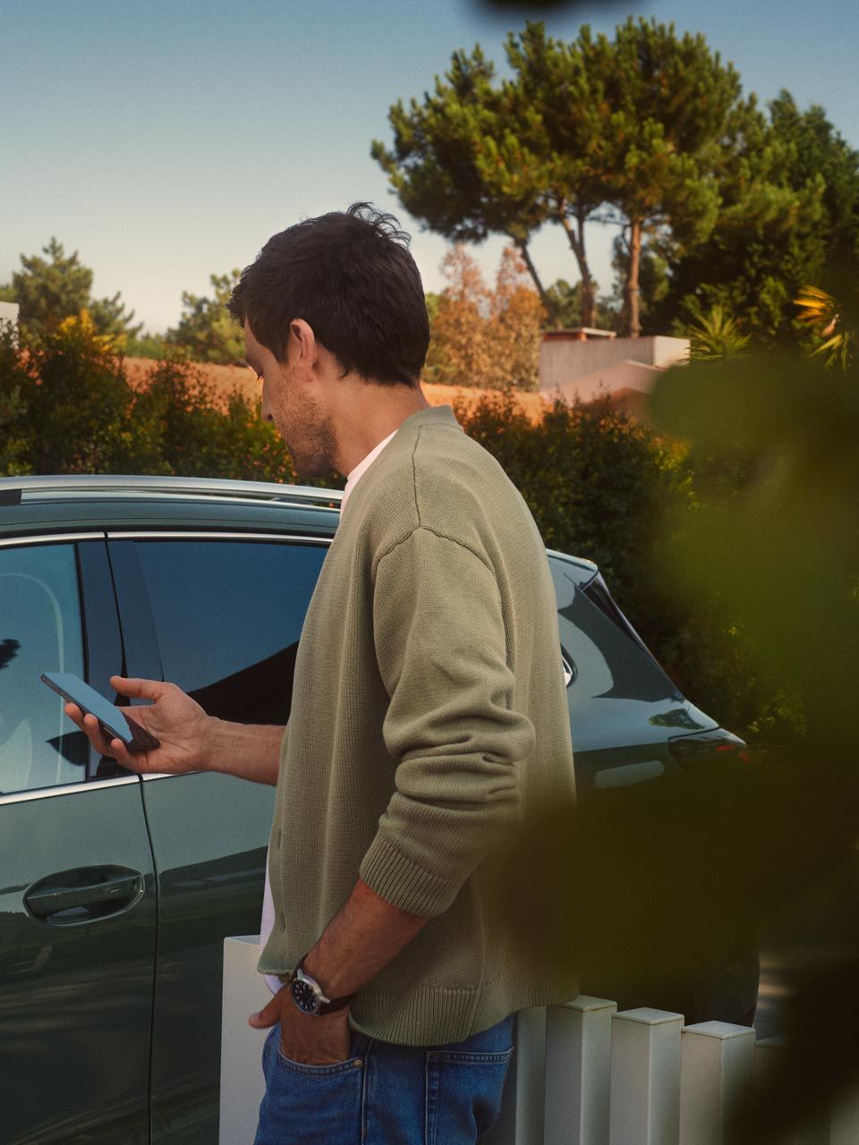 Ein junger Mann auf der Fahrerseite des dunkelgrünen VW Tiguan bedient den optionalen Park Assist Plus über sein Smartphone.