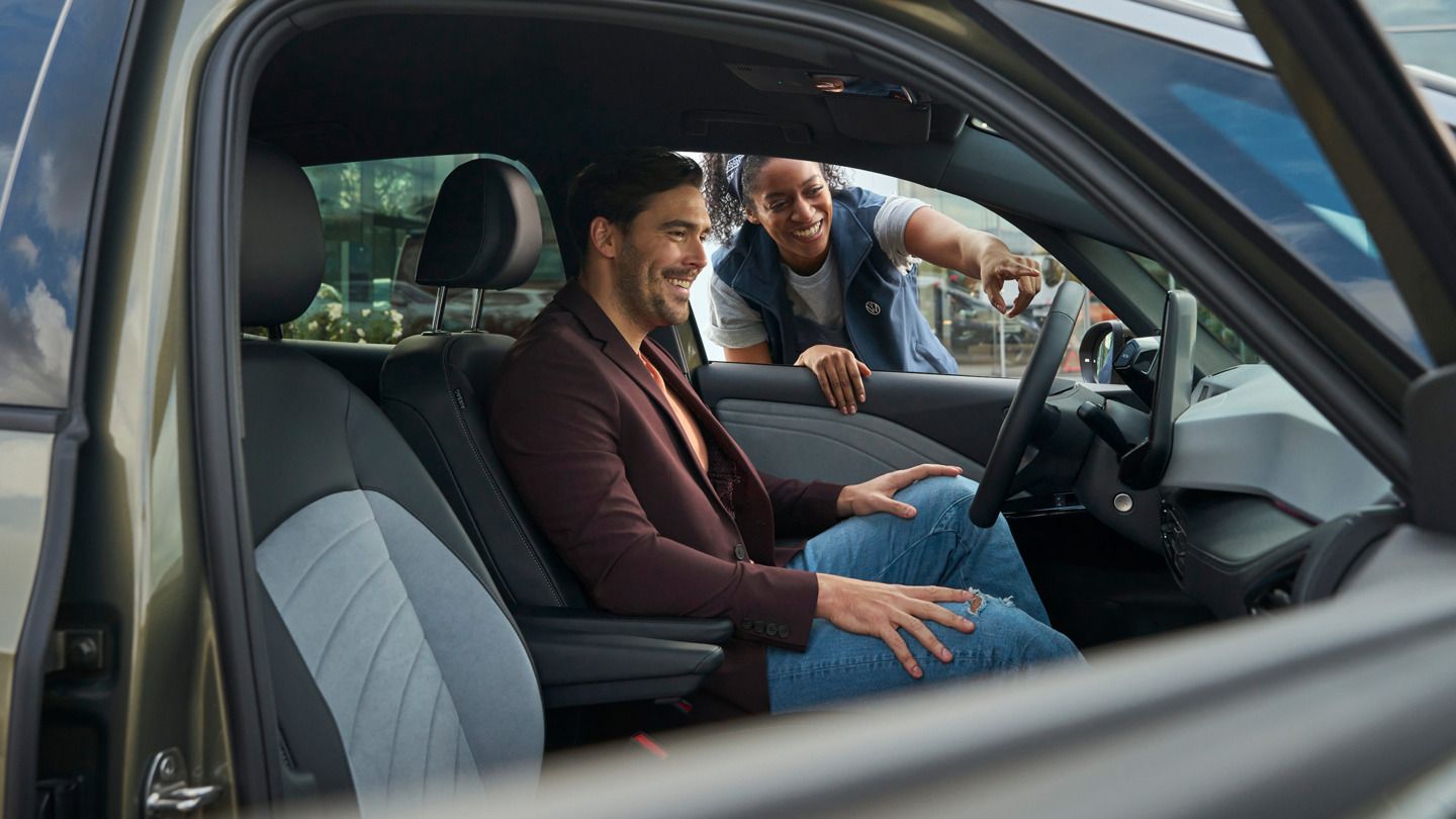 Egy férfi ül egy Volkswagen ID vezetőülésén, egy nő kívülről behajol a járműbe, és a műszerfalra mutat.