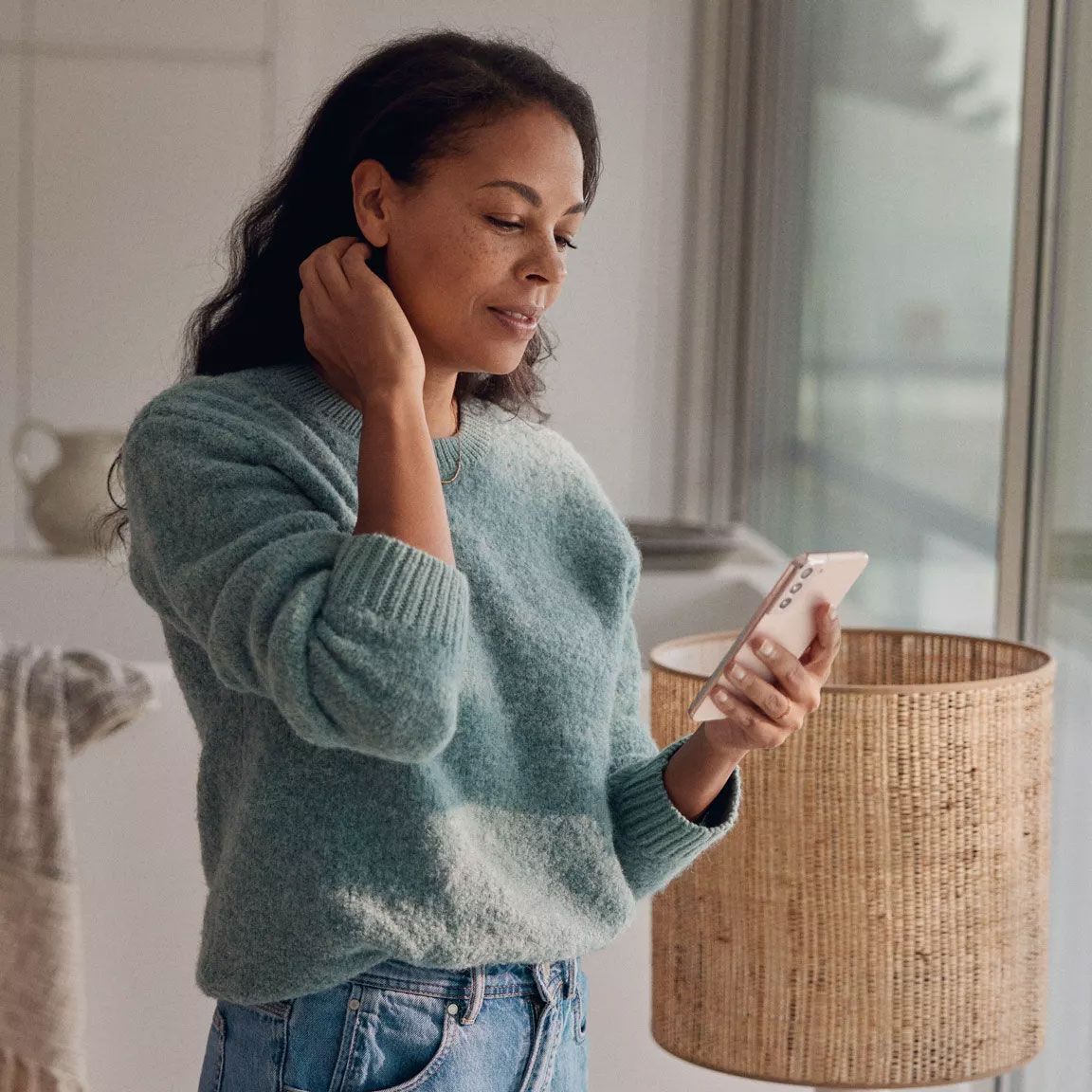 Egy nő otthon áll az ablaka előtt, és a mobiltelefonját nézi.