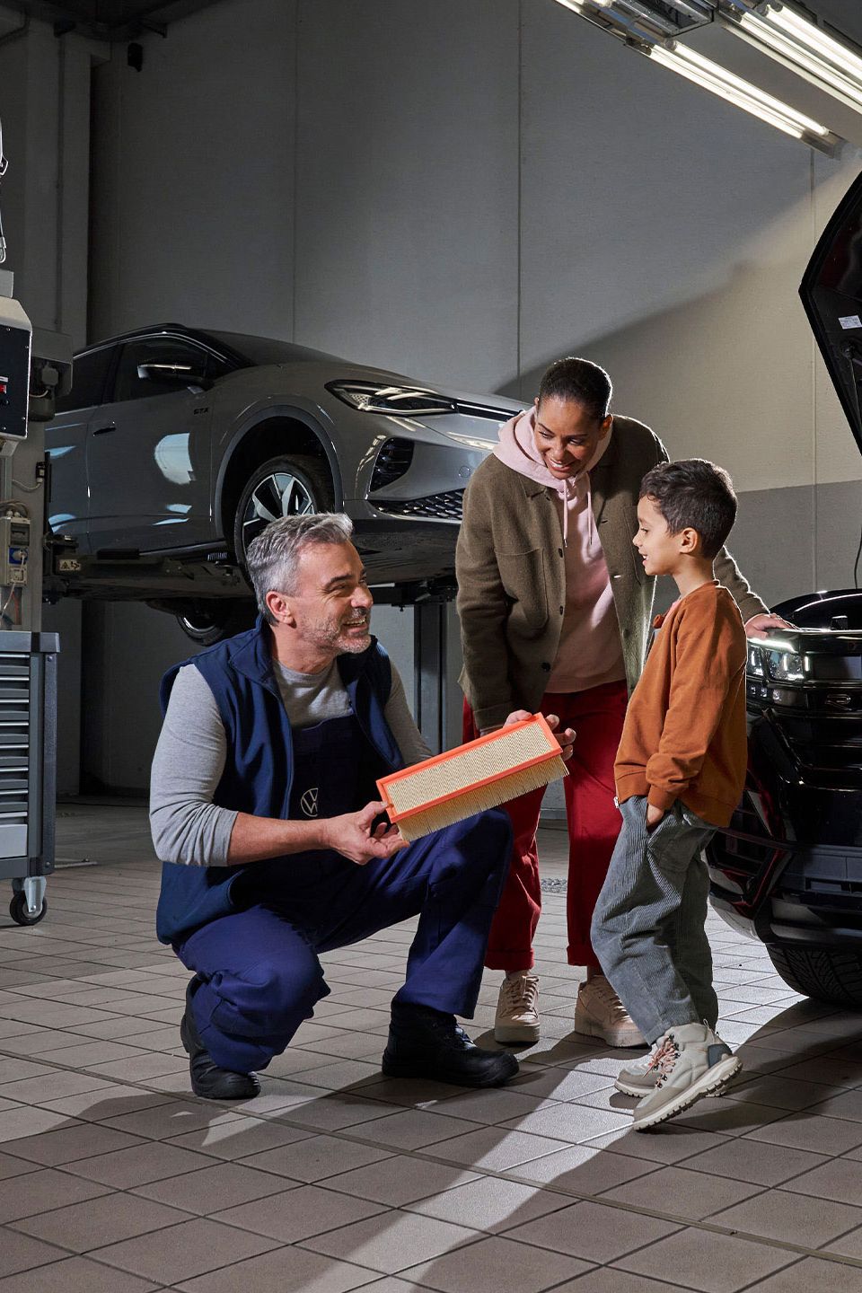 Ein Mechaniker erklärt den Aufbau eines Filters in einer Autowerkstatt. Im Hintergrund sind ein angehobenes Auto und ein geöffneter Motorraum eines schwarzen Touareg