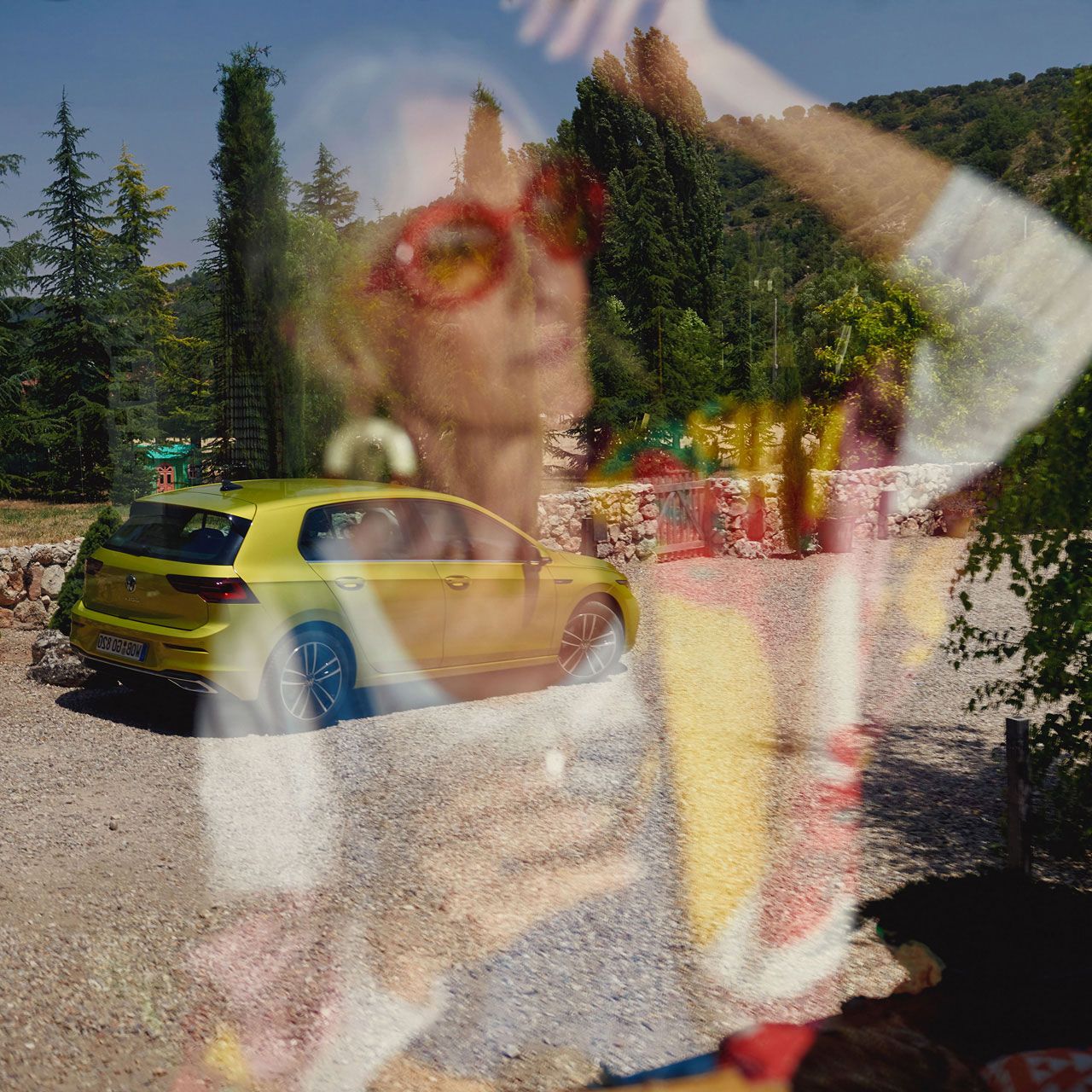 VW Golf in Gelb parkt seitlich vor Bäumen. Spiegelbild einer Frau ist im Vordergrund verschwommen zu sehen.