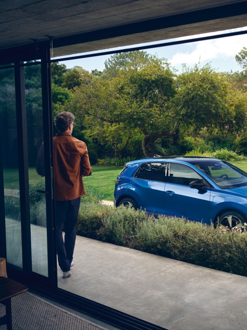 Blick durch eine offene Terrassentür in einen grünen Garten, in der Auffahrt steht ein blauer VW ID. Polo Style.