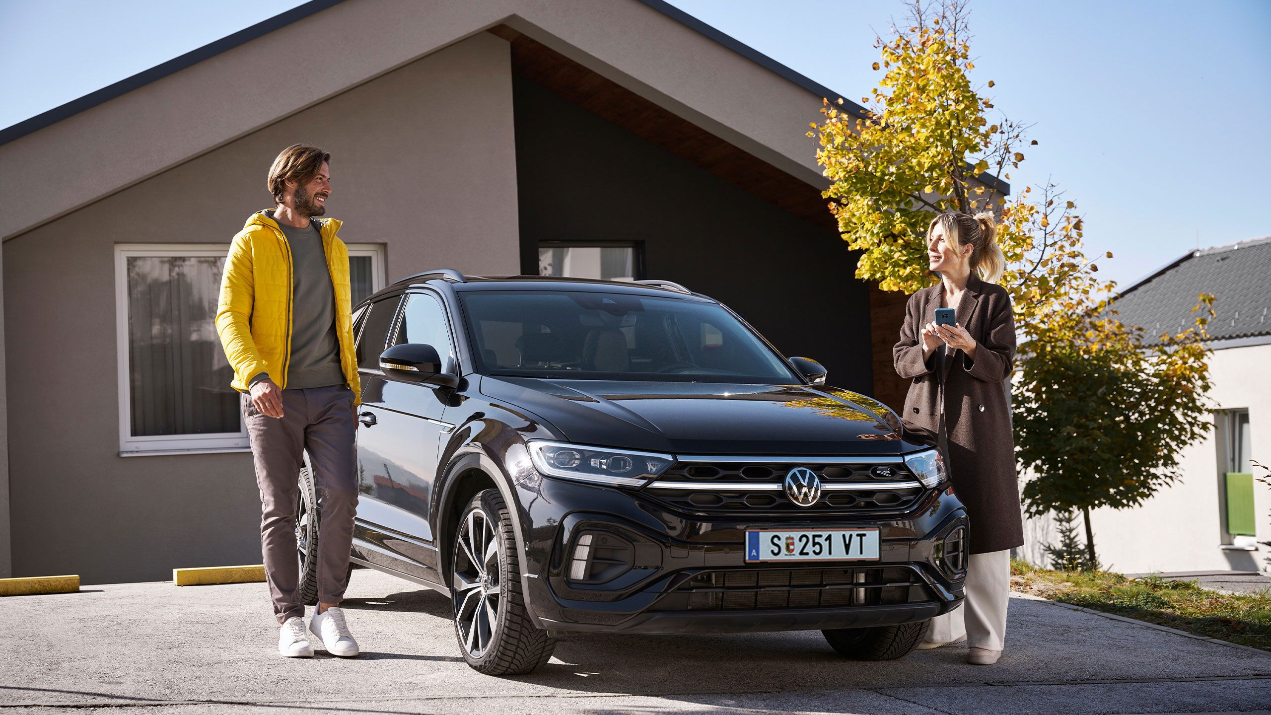 Egy férfi és egy nő mobiltelefonnal állnak egy Volkswagen előtt egy lakóépület előtt.