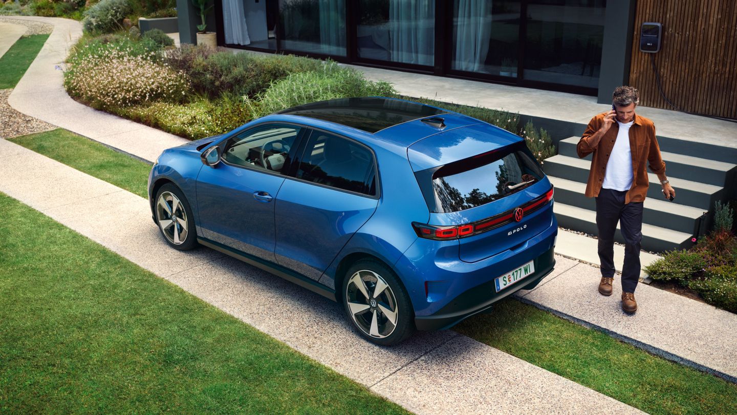Ein blauer VW ID. Polo Style parkt vor einem begrünten Wohnhaus, ein Mann geht hinter dem Fahrzeug entlang.