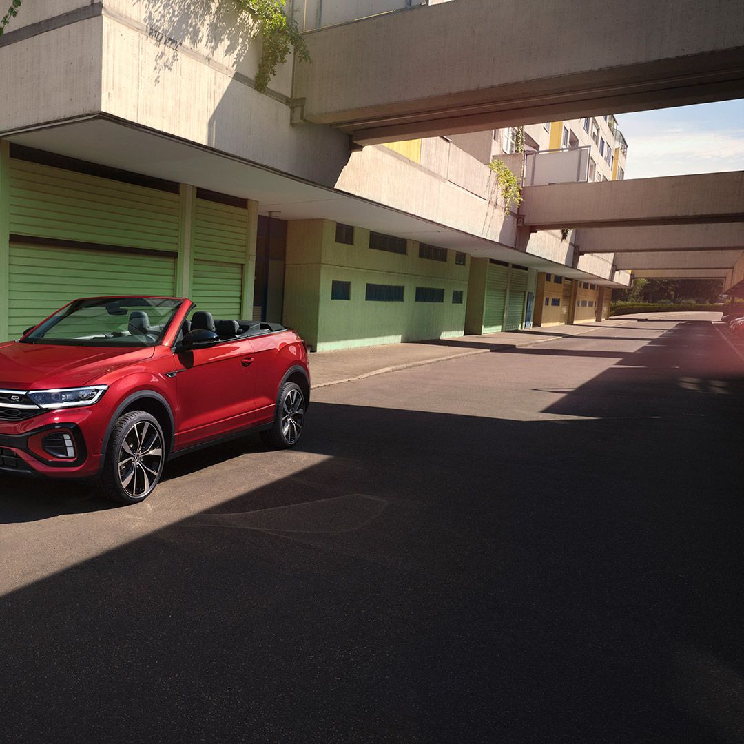Rotes VW T-Roc Cabriolet mit geöffnetem Dach parkt am Straßenrand. Eine Frau mit zwei Hunden steht daneben. 